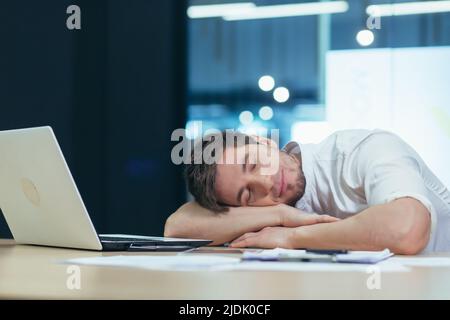 Il giovane bel freelance, uomo d'affari, direttore in ufficio al tavolo di riposo, si addormentò fra le braccia al tavolo, stanco Foto Stock