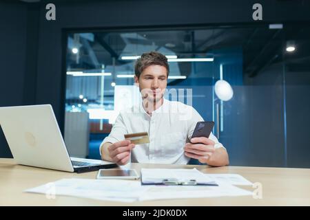 Un giovane è seduto in ufficio alla sua scrivania, con una carta di credito e un telefono cellulare, lavorando su un computer portatile. Effettua ordini online, paga le fatture Foto Stock