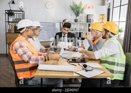 Gruppo diversificato di architetti, designer e ingegneri seduti al tavolo con gadget e progetti moderni di fronte a un enorme schermo TV al plasma. Uomini d'affari che lavorano su un progetto di costruzione comune. Foto Stock