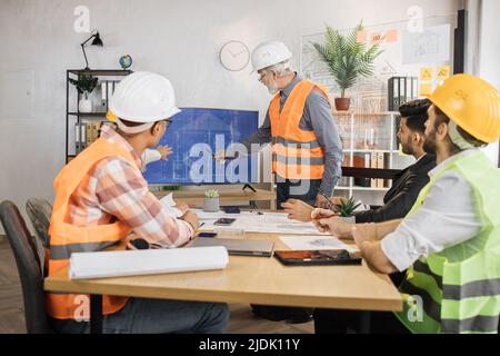 Uomo anziano in uniforme e casco che presenta un nuovo progetto di costruzione ai colleghi sullo schermo tv in ufficio. Tre uomini e una donna seduti a tavola e ascoltando il loro partner competente. Foto Stock