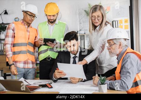 Designer creativi, architetti e ingegneri che si riuniscono in uffici moderni per pianificare nuovi progetti di costruzione. Gruppo di cinque persone multietniche che cooperano. Foto Stock