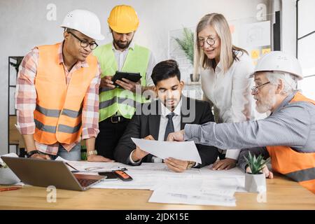 Designer creativi, architetti e ingegneri che si riuniscono in uffici moderni per pianificare nuovi progetti di costruzione. Gruppo di cinque persone multietniche che cooperano. Foto Stock