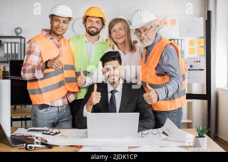 Team di architetti, ingegneri e designer qualificati che si incontrano nella sala riunioni dell'ufficio al coperto, in piedi vicino al boss indiano, seduti sulla scrivania con il computer portatile, guardando la fotocamera. Foto Stock