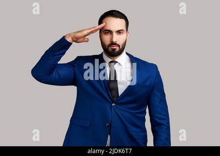 Ritratto di grave uomo barbuto stretto che tiene la mano vicino alla testa, salendo, onore e gloria, militarizzazione, indossando vestito di stile ufficiale. Studio interno girato isolato su sfondo grigio. Foto Stock