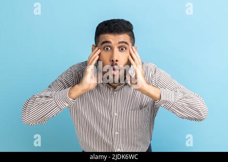 WOW, è incredibile. Sorprende l'uomo d'affari che fissava la macchina fotografica con la bocca aperta e gli occhi grandi, sollevando le braccia con stupore, indossando una camicia a righe. Studio interno girato isolato su sfondo blu. Foto Stock