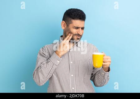 Ritratto di un uomo d'affari travagliato che soffre di un terribile dolore ai denti dopo aver bevuto bevande calde o fredde, lesioni dentali, indossando una camicia a righe. Studio interno girato isolato su sfondo blu. Foto Stock