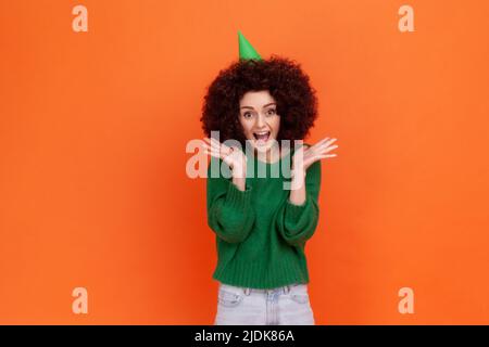 Ritratto di donna eccitata con acconciatura Afro indossando il maglione verde casual stile e cono festa, festeggiare il compleanno, urlando, felice di sorpresa. Studio interno girato isolato su sfondo arancione. Foto Stock