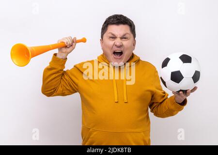Molto felice allegro uomo di mezza età che soffia in grande corno che tiene la palla di calcio, celebrando l'inizio del campionato, indossando felpa con cappuccio in stile urbano. Studio interno girato isolato su sfondo bianco. Foto Stock