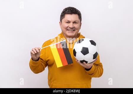 Uomo di mezza età con il sorriso toothy che tiene la bandiera della germania e calcio palla classica in bianco e nero e guardare la partita, indossando felpa con cappuccio urban style. Studio interno girato isolato su sfondo bianco. Foto Stock