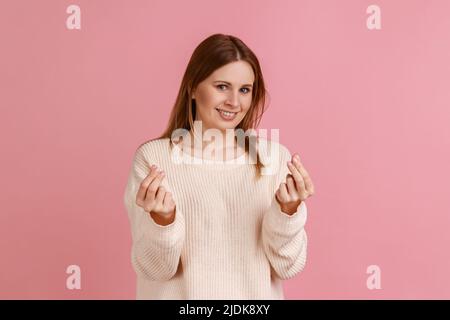 Ritratto di bella donna bionda positiva sorridendo e mostrando mi dare denaro gesto, chiedendo pagamento, indennità, indossare maglione bianco. Studio interno girato isolato su sfondo rosa. Foto Stock