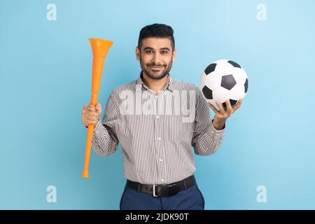 Un uomo d'affari con le bacchette che si batte per la squadra di calcio preferita, tenendo il corno e la palla in mano, guardando la macchina fotografica sorridendo, indossando una maglietta a righe. Studio interno girato isolato su sfondo blu. Foto Stock