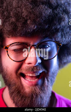 Ritratto closeup di un hipster positivo con acconciatura afro di buon umore, sorridendo ampiamente e sverchiando alla macchina fotografica, mantiene la bocca aperta. Studio interno girato isolato su colorato sfondo di luce al neon. Foto Stock