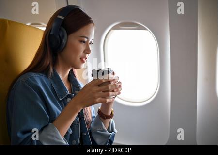 Attraente donna asiatica millenaria con cuffie rilassa sorseggiando un caffè caldo su un volo. Viaggio, vacanza, concetto di turismo Foto Stock