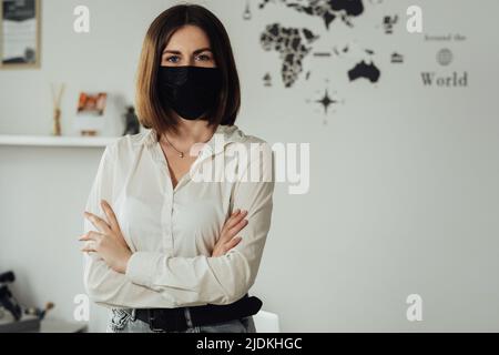 Ritratto di giovane donna caucasica indossare maschera di protezione in piedi in ufficio con le braccia di tenuta, spazio copia Foto Stock