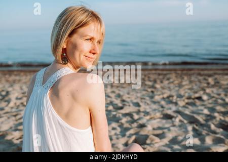 Vista posteriore di una donna sorridente di mezza età seduta sulla spiaggia di sabbia vicino mare mare oceano in giorno di sole, guardando indietro. Estate. Foto Stock