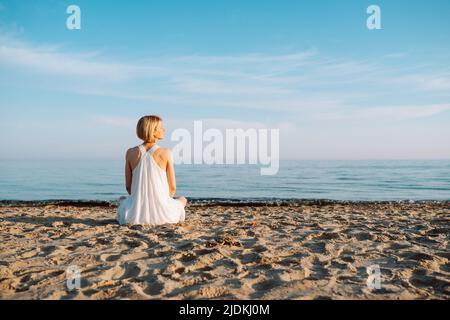 Vista posteriore di donna sottile indossando bianco luce estate Sundress seduta sulla spiaggia di sabbia vicino mare mare oceano in giorno di sole. Foto Stock