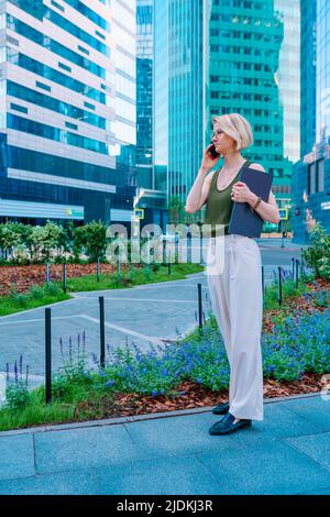 Vista laterale della donna bionda in piedi sulla strada nel centro della città vicino grattacieli, tenendo un computer portatile blu, parlando sullo smartphone. Foto Stock