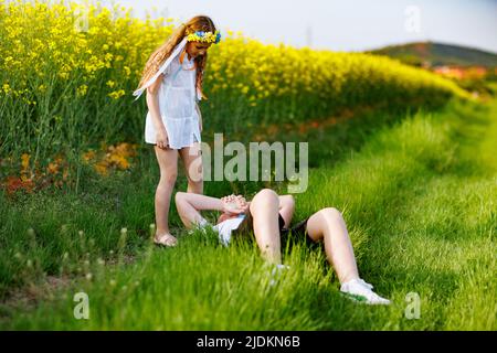 Allegra ragazza sorridente gioiosa con brillante corona di fiori ucraini con lunghi nastri multicolore si alza e guarda il fratello adolescente, su cui si trova Foto Stock