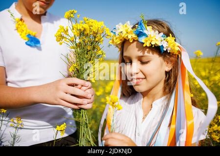 Allegro gioiosa bambini spensierati: Fratello e sorella in corona fiorita Ucraina con nastri multicolore fiutano fiori di colza selvaggia in mazzi aga Foto Stock