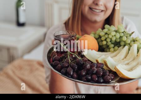 Inriconoscibile giovane donna che tiene vassoio di frutta in camera d'albergo, bottiglia di vino si trova sullo sfondo Foto Stock