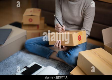 Giovane donna piccola impresa proprietario online shopping a casa. Confermare gli ordini da parte dei clienti con il telefono cellulare preparare il prodotto pacchetto in background. PMI Foto Stock