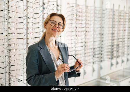 La donna sceglie tra due cornici da spettacolo nel negozio di un ottico e guarda la fotocamera Foto Stock