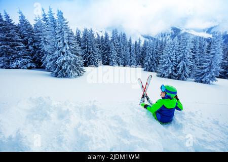 Ragazzo si siede nella neve con sci sopra la foresta di abeti dopo nevicate pesanti Foto Stock