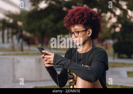 vista laterale, bella giovane donna bruna latina con afro, sorridente felice seduta all'aperto controllare il suo telefono, controllare le notifiche sul social network Foto Stock