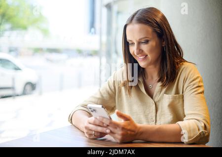 Giovane donna che usa lo smartphone seduto alla scrivania in un bar, sorridente dipendente che chiacchiera online durante la pausa. La signora allegra ama inviare SMS online, usa app mobile Foto Stock