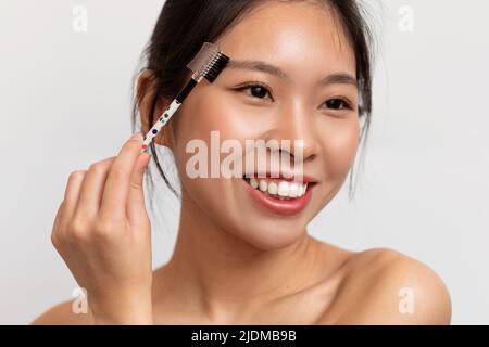 Bella signora coreana spazzolando le sopracciglia e applicando mascara sopracciglia, utilizzando trucco decorativo, sfondo bianco Foto Stock