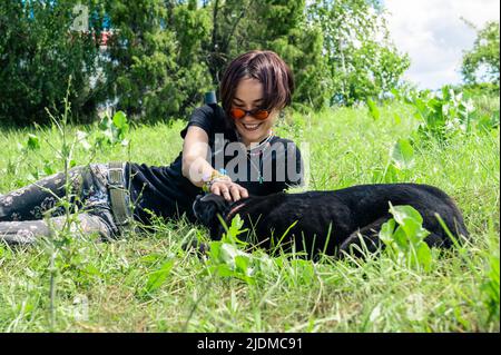 Bella ragazza felice giace e gioca sul prato con il suo cane. Giovane donna che coccola il suo cane sull'erba. Concetto di amicizia Foto Stock