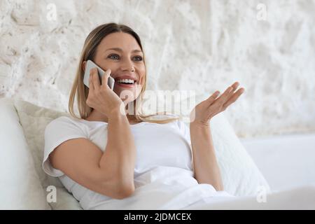 Bella donna bionda seduta a letto, parlando al telefono Foto Stock