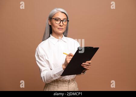 Donna d'affari senior fiduciosa che scrive negli appunti e guarda la fotocamera. Dipendente femminile maturo in abbigliamento formale isolato su sfondo beige Foto Stock