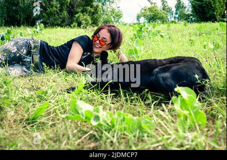 Bella ragazza felice giace e gioca sul prato con il suo cane. Giovane donna che coccola il suo cane sull'erba. Concetto di amicizia Foto Stock