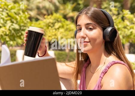 Giovane donna seduta in un parco che tiene un bicchiere di caffè in una mano e un libro nell'altra mentre ascolta la musica sulle cuffie. Foto Stock