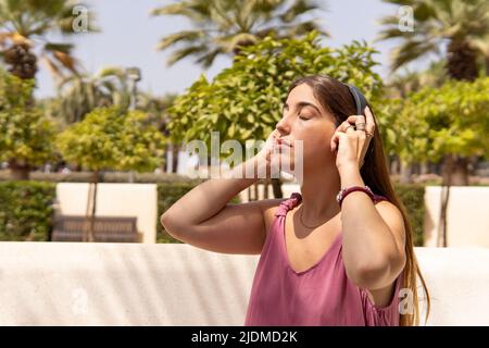 Bella ragazza in abito rosa seduta su una panca parco ascoltare musica, tenendo cuffie con entrambe le mani, sentire la canzone. Foto Stock