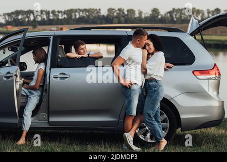 Uomo e donna caucasica che abbraccia vicino alla Minivan Car, Happy Four Members Family godersi il viaggio, facendo sosta presso il fiume Foto Stock