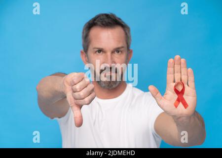 Uomo maturo che gestizza il pollice con un fiocco rosso simbolo di consapevolezza AIDS su una palma che indossa una maglietta bianca isolata su sfondo blu. Medicina moderna e assistenza sanitaria. CONCETTO DI SENSIBILIZZAZIONE DELL'AIDS. Foto Stock