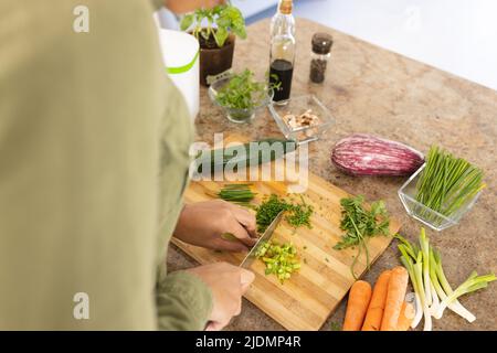 Sezione centrale della giovane donna biraciale che trita verdure sul tagliere in cucina a casa Foto Stock