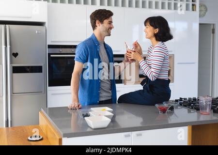 Felice coppia giovane multirazziale tenendo frullato in vetro guardando l'un l'altro in cucina Foto Stock