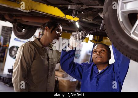 Meccanica femminile asiatica che aiuta il collega di adulti medio afroamericano a riparare l'auto in officina Foto Stock