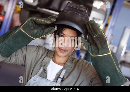 Ritratto di sicura saldatrice femminile asiatica di metà adulto indossando indumenti da lavoro protettivi in officina Foto Stock