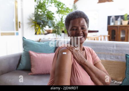 Ritratto di sorridente anziana afroamericana con bendaggio adesivo sulla spalla seduta sul divano Foto Stock