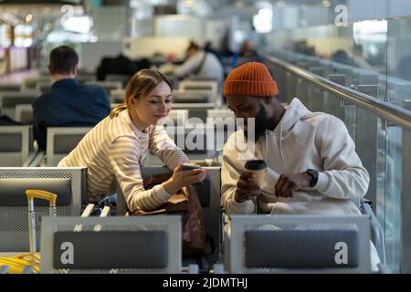 Maschio hipster nero e femmina caucasica bella guardando il telefono mentre si aspetta il volo in aeroporto. Foto Stock