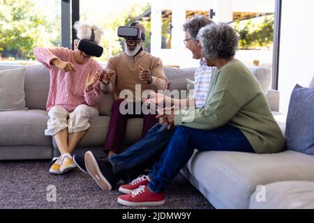 Anziani multirazziali che guardano amici allegri gesturando usando simulatori di realtà virtuale Foto Stock
