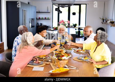 Felice multirazziale senior amici tostare wineglasses mentre si pranzano al tavolo da pranzo Foto Stock