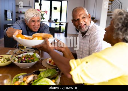 Sorridendo l'anziano biraciale che dà il pane alla donna mentre pranzano al tavolo da pranzo nella casa di cura Foto Stock