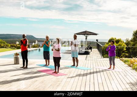 Gli anziani multirazziali meditano mentre si levano in piedi sul pavimento del legno duro a bordo piscina contro il cielo in estate Foto Stock