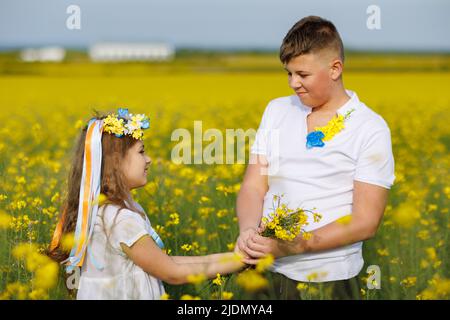 Allegro gioiosa bambini spensierati: Fratello e sorella in corona fiorita Ucraina con nastri multicolore fiutano fiori di colza selvaggia in mazzi aga Foto Stock