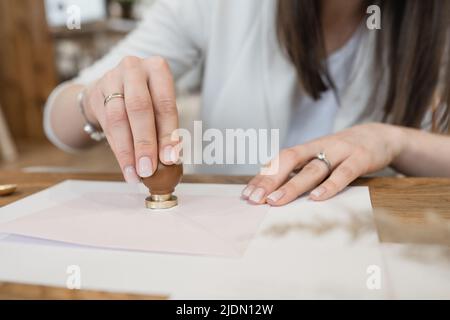 Mani ben curate con anelli di donna irriconoscibile che fanno sigillo di cera con timbro su busta bianca su scrivania di legno. Foto Stock